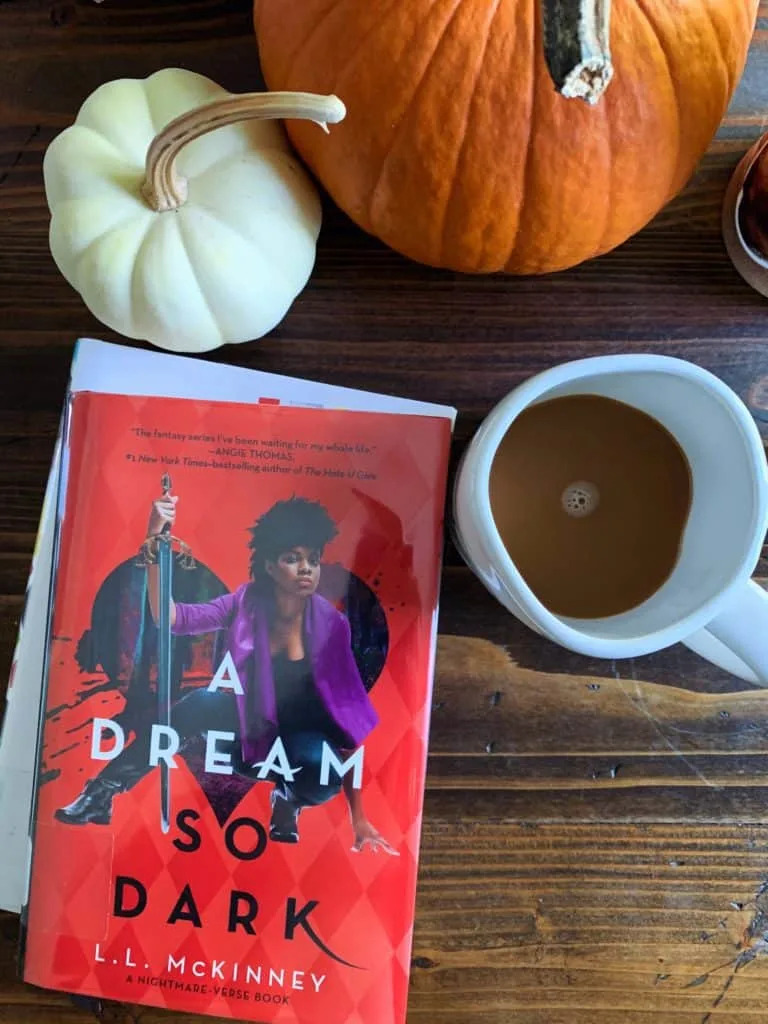 There are pumpkins coffee and a book A Dream So Dark on the table. 
